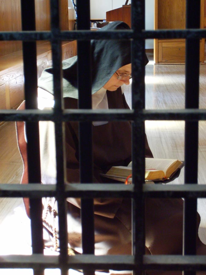 Das Photo zeigt eine Nonne im traditionellen Habit der Karmeliterinnen, die hinter dem Klausurgitter sitzend das Brevier betet.