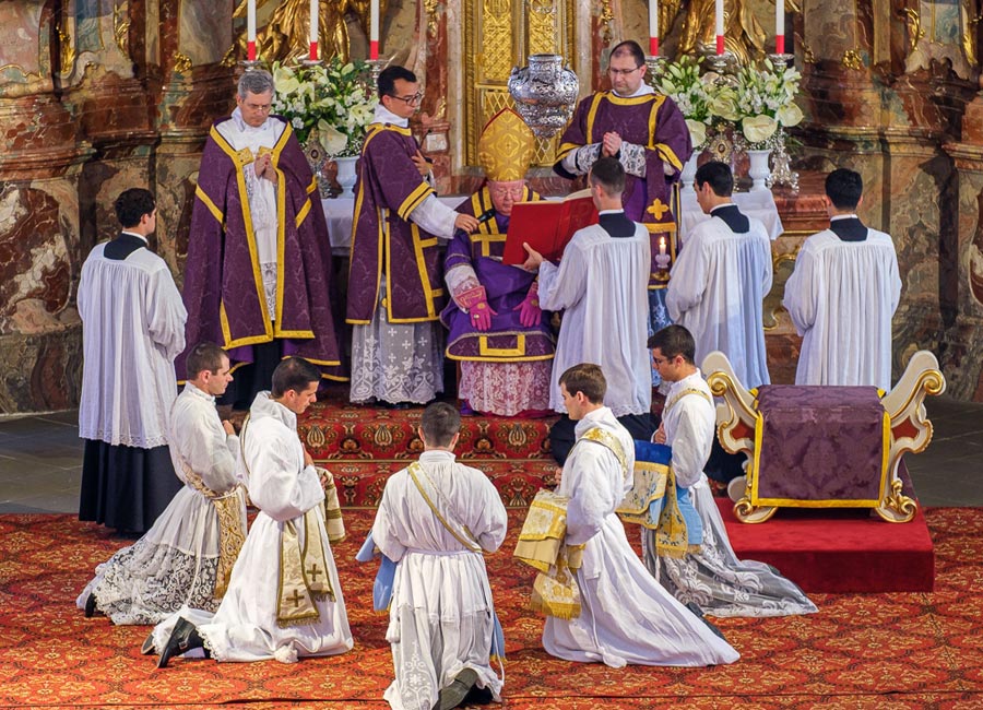 Der Bischof sitzt auf einem Schemel vor dem Altar - die Weihekandidaten knien im Halbkreis davor.