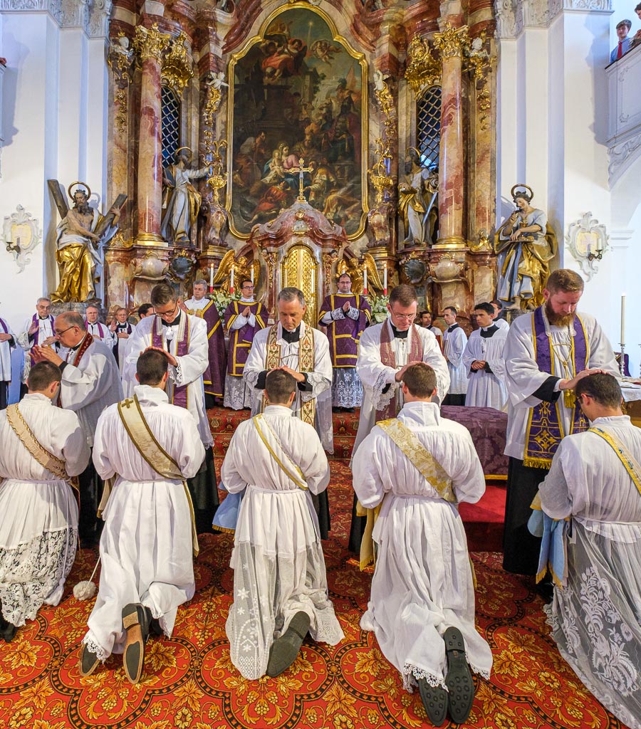 Die Neugeweihten knien in einer Reihe vor dem Altar, während alle anwesenden Priester ihnen ebenfalls die Hände auflegen.