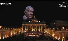 Das Bild zeigt den nächtlich dunklen Petersplatz mit der Veranstaltung und über dem Petersdom von hunderten Leucht-Drohnen dargestellt das gütig herballächelnde Gesicht eines überirdischen Franziskus